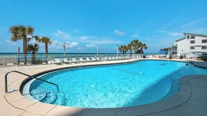 A pool with a curved edge, surrounded by lounge chairs and palm trees, with a view of the ocean and a building in the background. - Watercrest 1005 (Panama City Beach)
