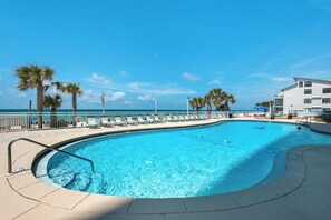 A pool with a curved edge, surrounded by lounge chairs and palm trees, with a view of the ocean and a building in the background. - Watercrest 1005 (Panama City Beach)