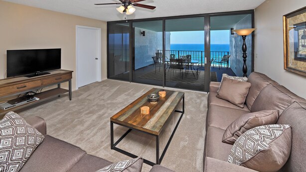 A living room with a TV, sofa, coffee table, and a balcony with a view of the ocean. - Watercrest 1005 (Panama City Beach)