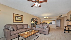 A living room with two sofas, a coffee table, a ceiling fan, a kitchen in the background, and a framed picture on the wall. - Watercrest 1005 (Panama City Beach)