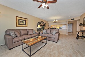 A living room with two sofas, a coffee table, a ceiling fan, a kitchen in the background, and a framed picture on the wall. - Watercrest 1005 (Panama City Beach)