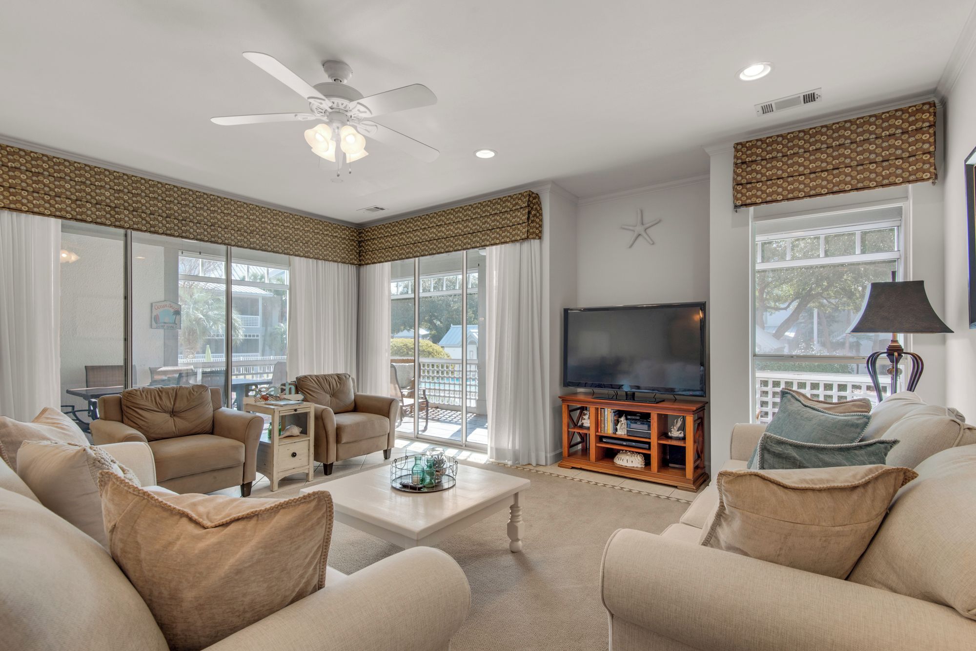 A spacious living room with a ceiling fan, large windows, a flat-screen TV, and a comfortable seating area.