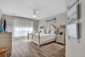 A bedroom with a bed, bedside tables, a ceiling fan, a window with curtains, and wall art. - Villa 18 at Hidden Dunes (Miramar Beach)