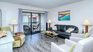 A modern living room with a glass coffee table, a black leather sofa, and a white armchair. There is a balcony with a view of the sea and a painting of a beach scene on the wall. - Runaway Bay Resort by BBG (Bradenton Beach)