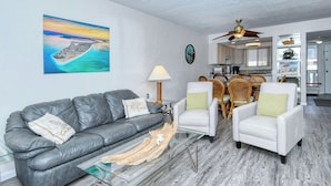 A living room with a gray leather sofa, white armchairs, a glass coffee table, and a wooden floor. - Runaway Bay Resort by BBG (Bradenton Beach)