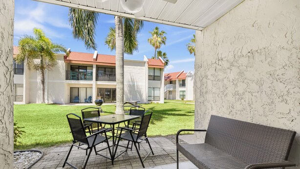Un patio techado con mesa y sillas, un sofá de mimbre y vistas a un edificio de techo rojo y palmeras.