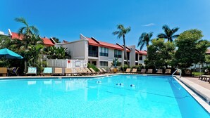 Una piscina con sillas de descanso y sombrillas, rodeada de palmeras y edificios con tejados rojos.