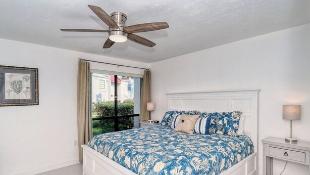 Ein Schlafzimmer mit einem Bett, einem Deckenventilator, einem Fenster mit Vorhängen, einem Nachttisch mit einer Lampe und einem gerahmten Bild an der Wand.