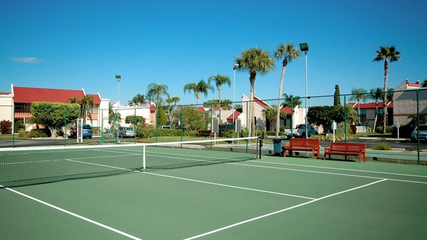 Een tennisbaan met een net, omringd door palmbomen en gebouwen met rode daken.