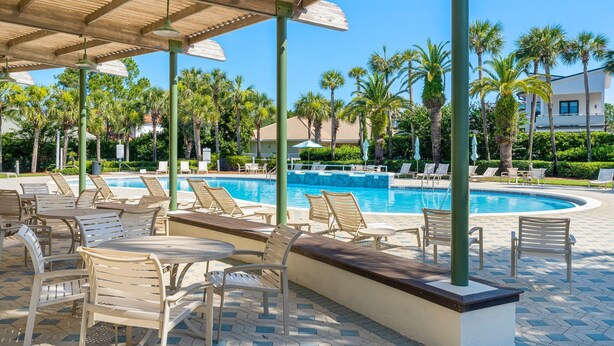 Zona junto a la piscina con sillones de descanso y sombrillas, rodeada de palmeras y un cielo azul despejado.