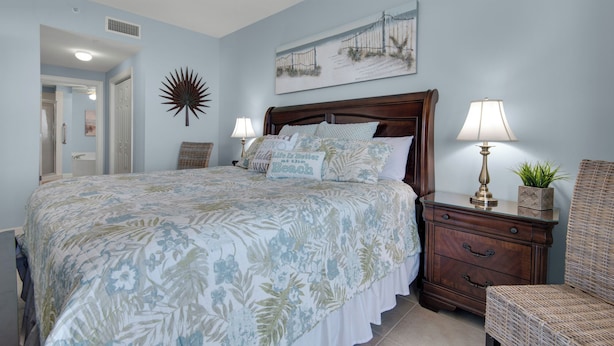 A bedroom with a large bed, two bedside tables with lamps, a chair, a decorative wall piece, and a door leading to another room.