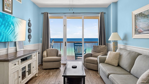 A living room with a sofa, chairs, a coffee table, a TV, and a view of the ocean through large sliding doors. - Sunrise Resort by BBG (Panama City Beach)