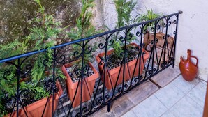 A narrow alley with a white brick wall and a stone wall on one side. There are potted plants along the wall and a metal railing in the foreground. - Alegria (Chania)
