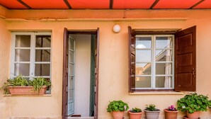 A building facade with two windows, one with a closed shutter and one with an open shutter. There are potted plants on the ground and a red awning above the windows. - Alegria (Chania)