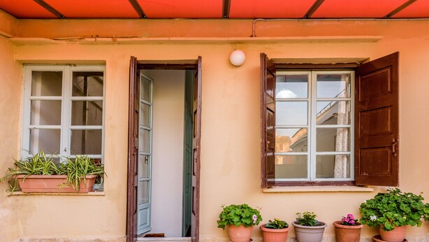 A building facade with two windows, one with a closed shutter and one with an open shutter. There are potted plants on the ground and a red awning above the windows. - Alegria (Chania)