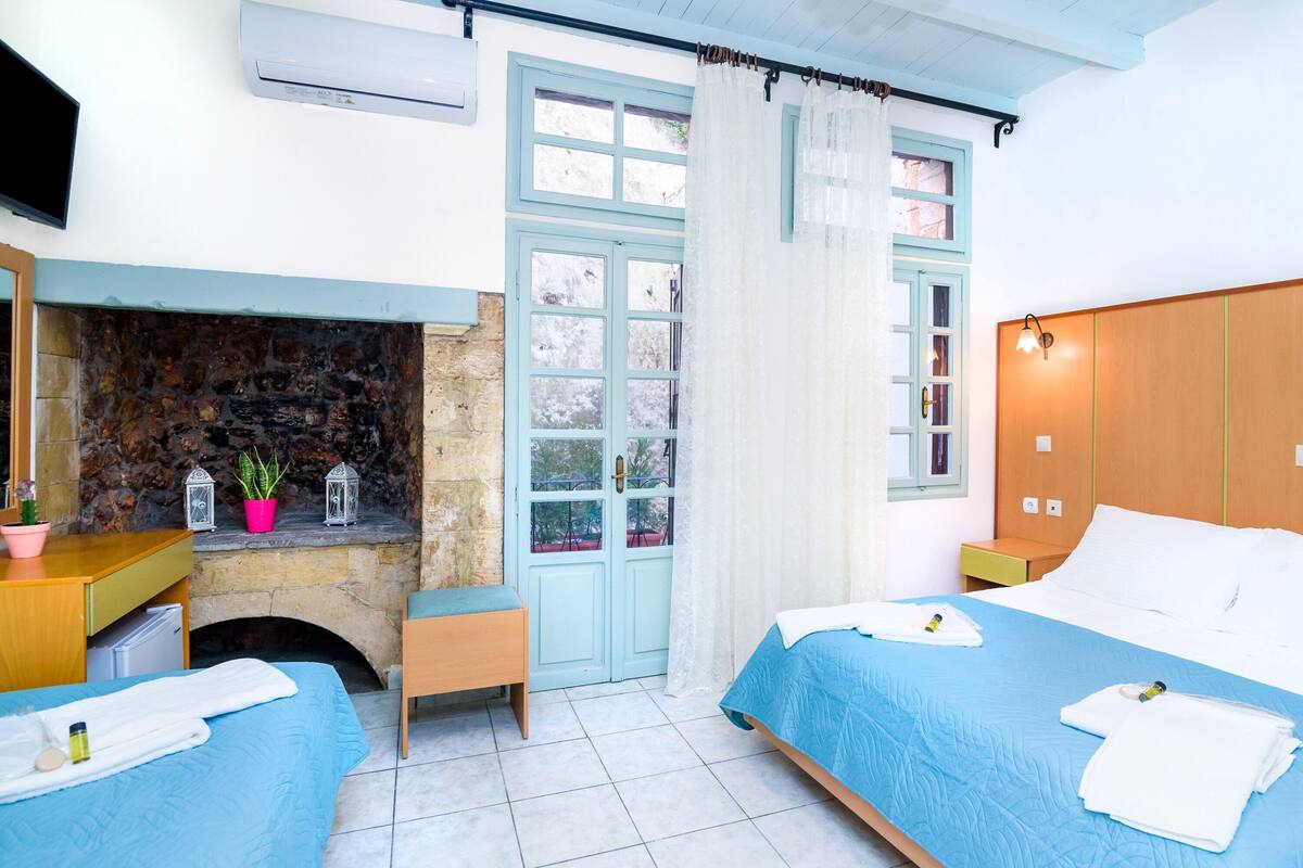 A bedroom with two beds, a stone fireplace, a window with curtains, and a nightstand with a lamp.