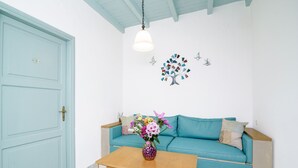 A room with a light blue door, a teal sofa, a wooden coffee table with a vase of flowers, and a wall-mounted light fixture. - Alegria (Chania)