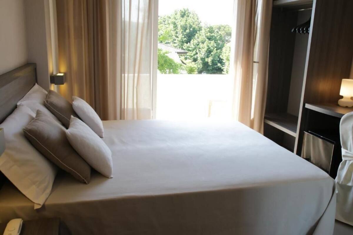A hotel room with a large bed, a bedside table with a lamp, and a view of greenery outside.