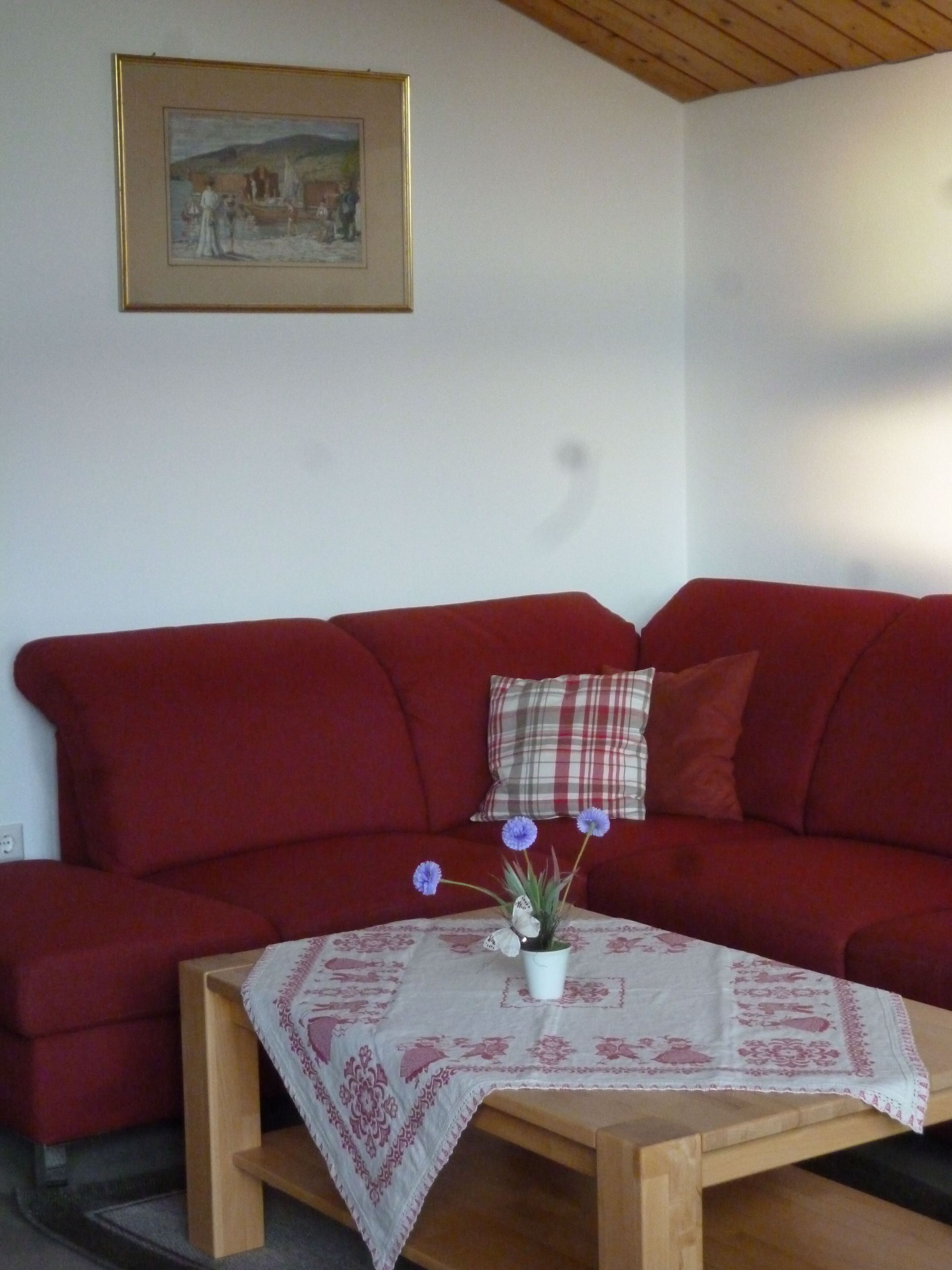 Un sofá rojo con un cojín escocés, una mesita de madera con mantel floral y un cuadro en la pared.