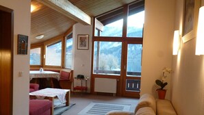 Ein gemütliches Wohnzimmer mit Holzbalkendecke, einer Couch, einem Tisch mit rotem Kissen und einem Fenster mit Blick auf die Berge.