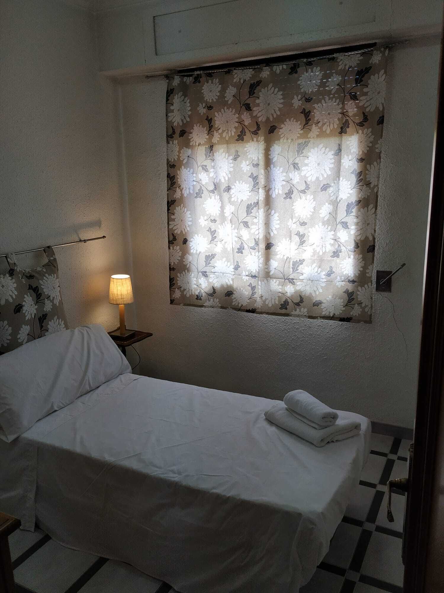 Een slaapkamer met een bed, een nachtkastje met een lamp, een raam met bloemen gordijnen, en een geruite vloer.