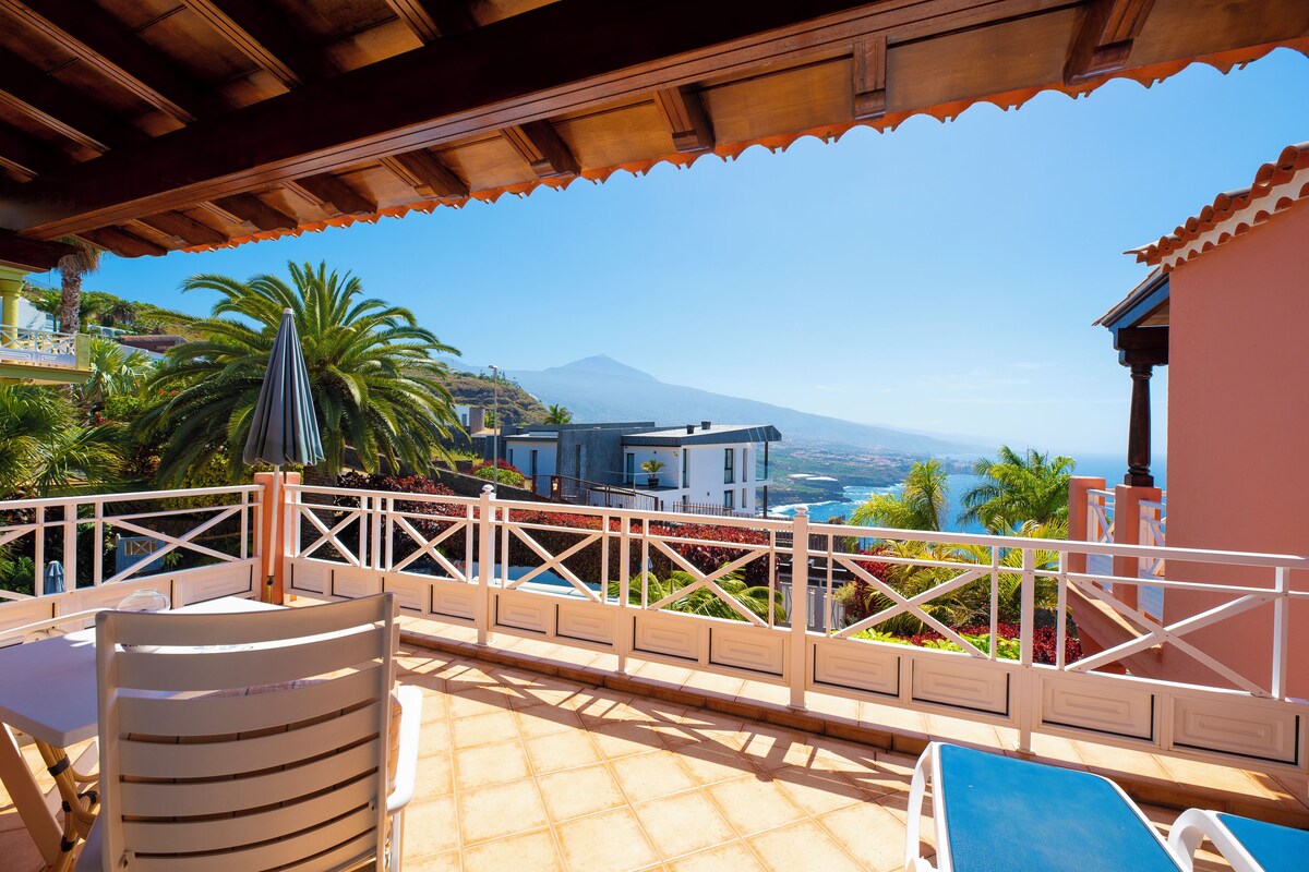 Une terrasse offrant une vue sur la montagne et la mer, avec une table, des chaises et une rambarde.