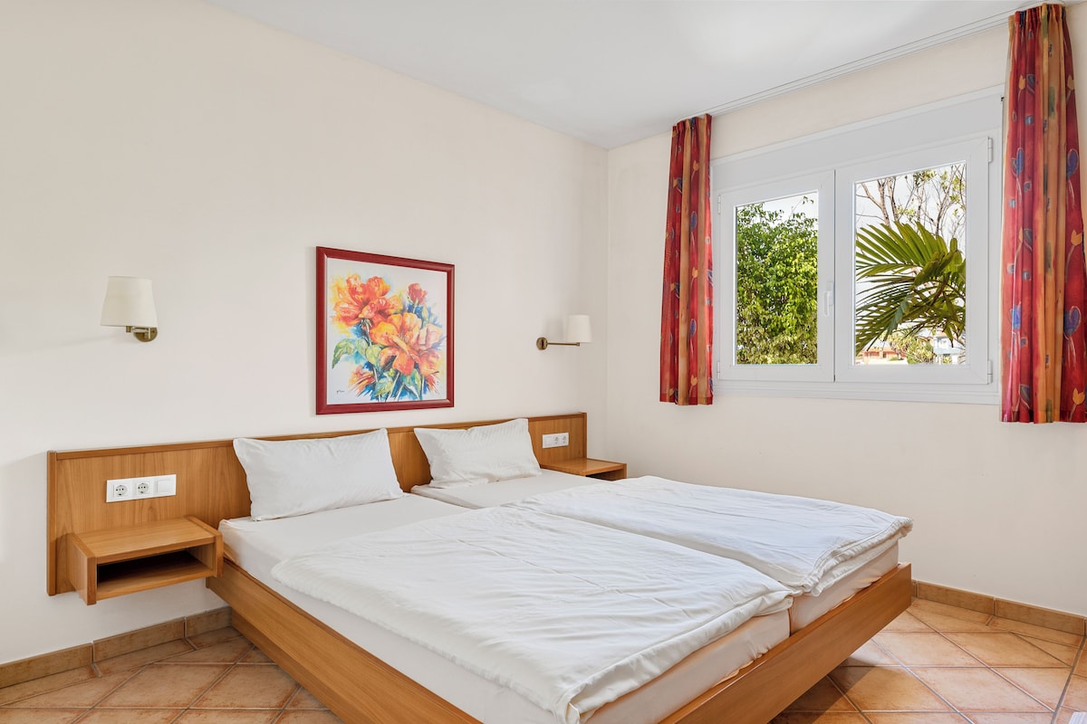 A double bed with a wooden headboard, two bedside tables, a wall-mounted lamp, a framed floral painting, and a window with curtains.