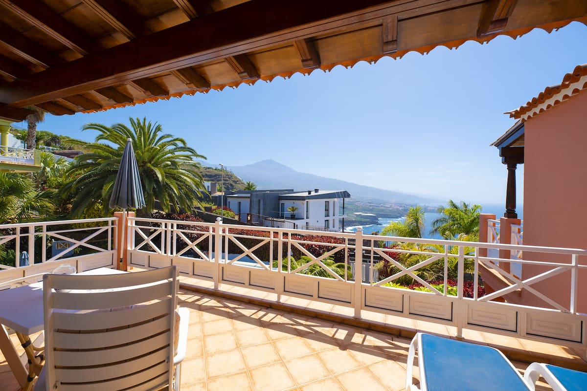 Eine Terrasse mit Blick auf einen Berg und das Meer, ausgestattet mit einem Tisch, Stühlen und einer Brüstung.