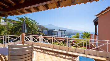 A terrace with a view of a mountain and the sea, featuring a table, chairs, and a railing.