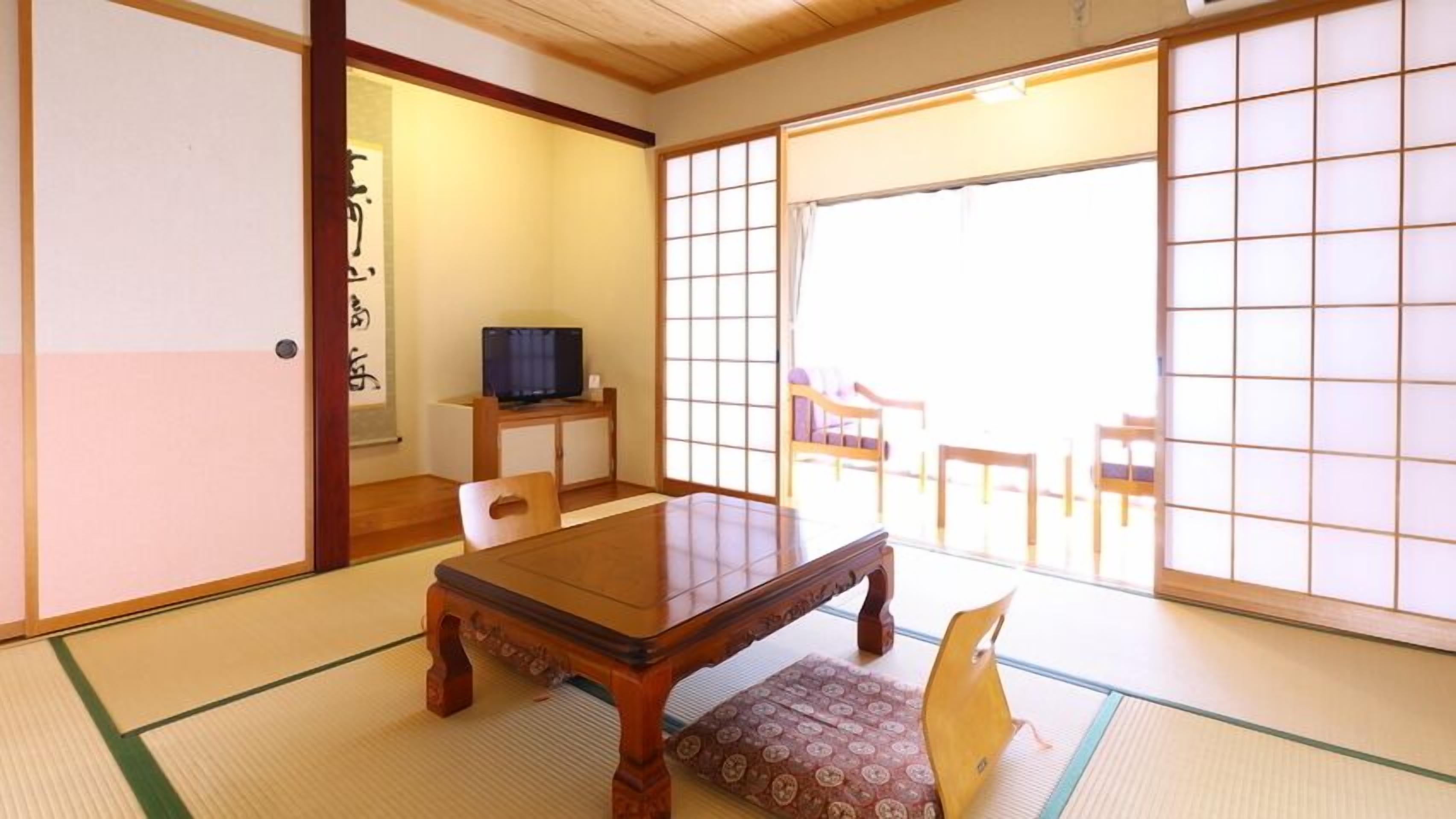 Habitació japonesa tradicional amb sòls de tatami, una taula de fusta, una televisió i portes corredisses que donen a un balcó.