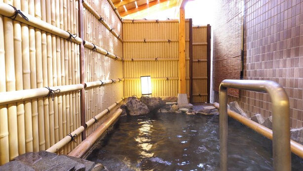 Un onsen japonés tradicional con paredes de bambú, una fuente termal de piedra y un pasillo de madera.