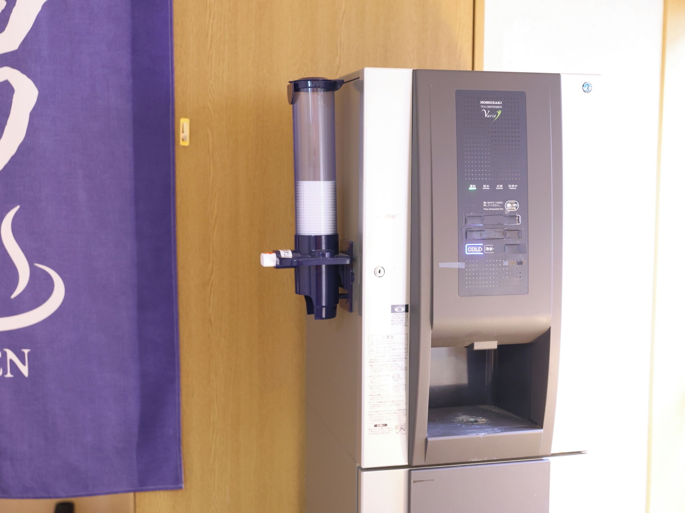 Un distributeur automatique équipé d’un point d’eau et d’une machine à café, fixé au mur à côté d’une porte en bois.