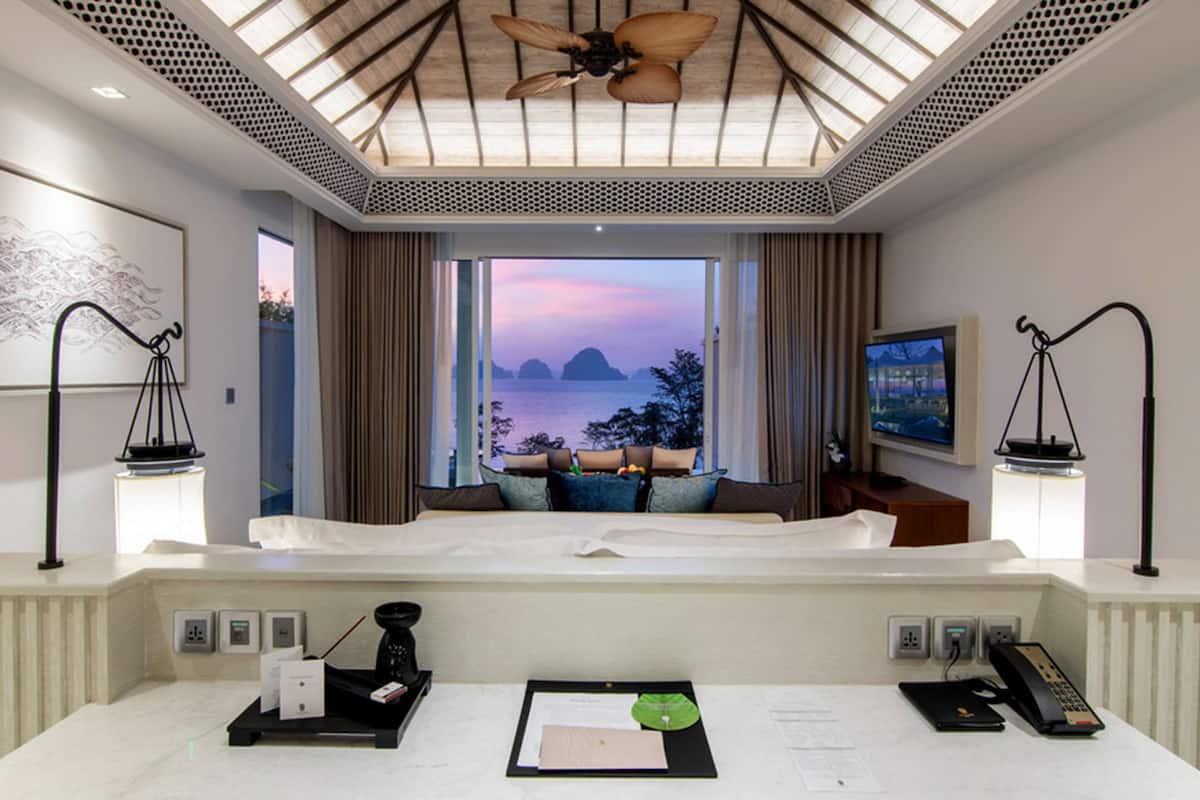 A hotel room with a bed, a desk with a computer, a telephone, and a view of the sea at sunset.