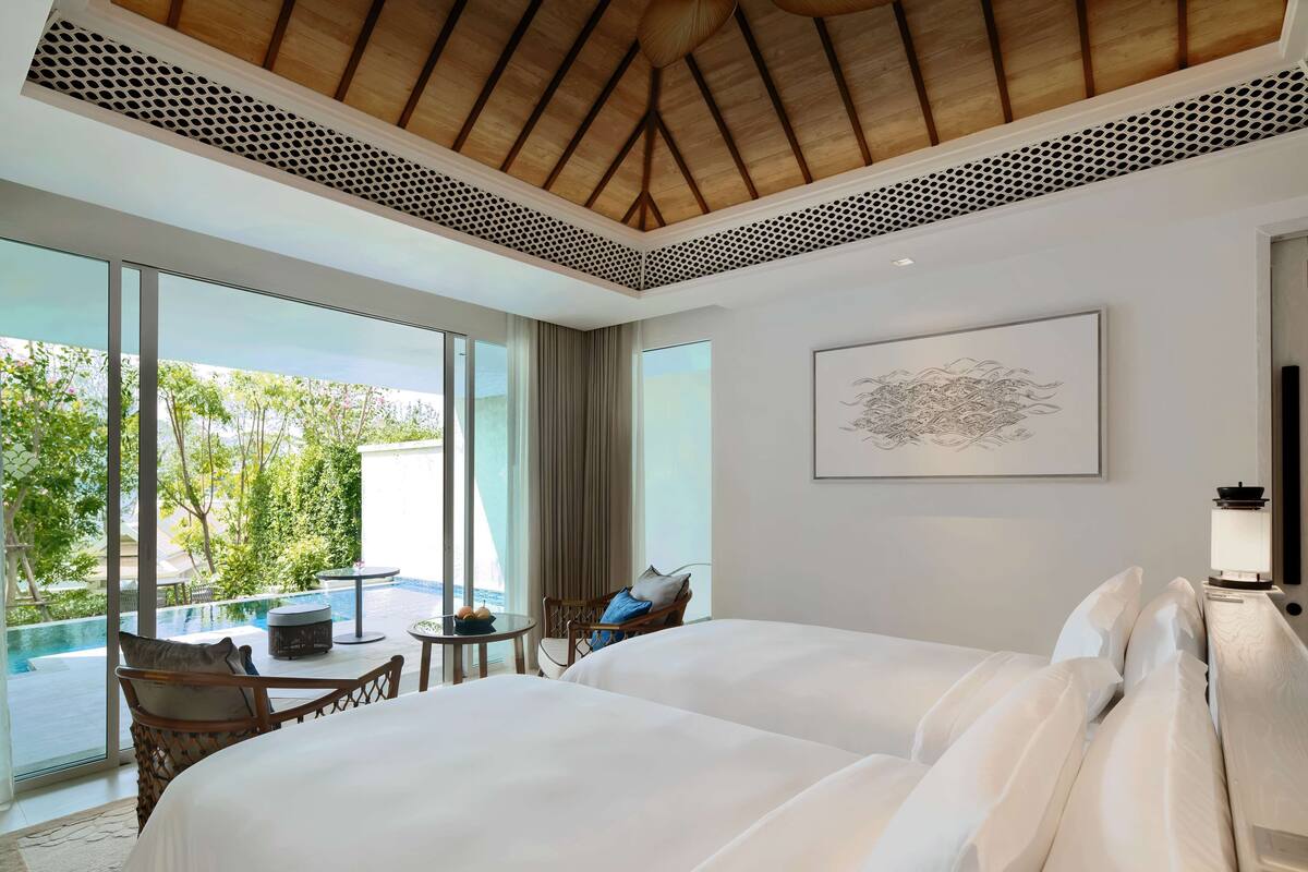 A hotel room with a large bed, a small table, a chair, and a view of a swimming pool and greenery.