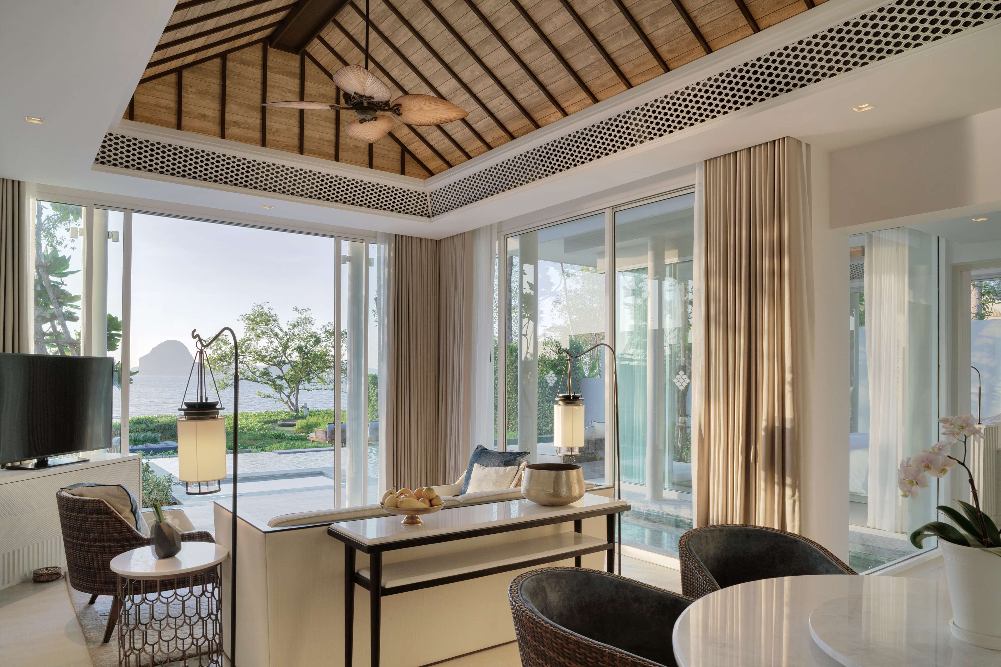 A modern hotel room with a large window, a ceiling fan, a flat-screen TV, and a dining area with a table and chairs.