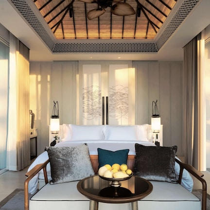 A modern hotel room with a large bed, a small round table with fruit, and a view of the pool.