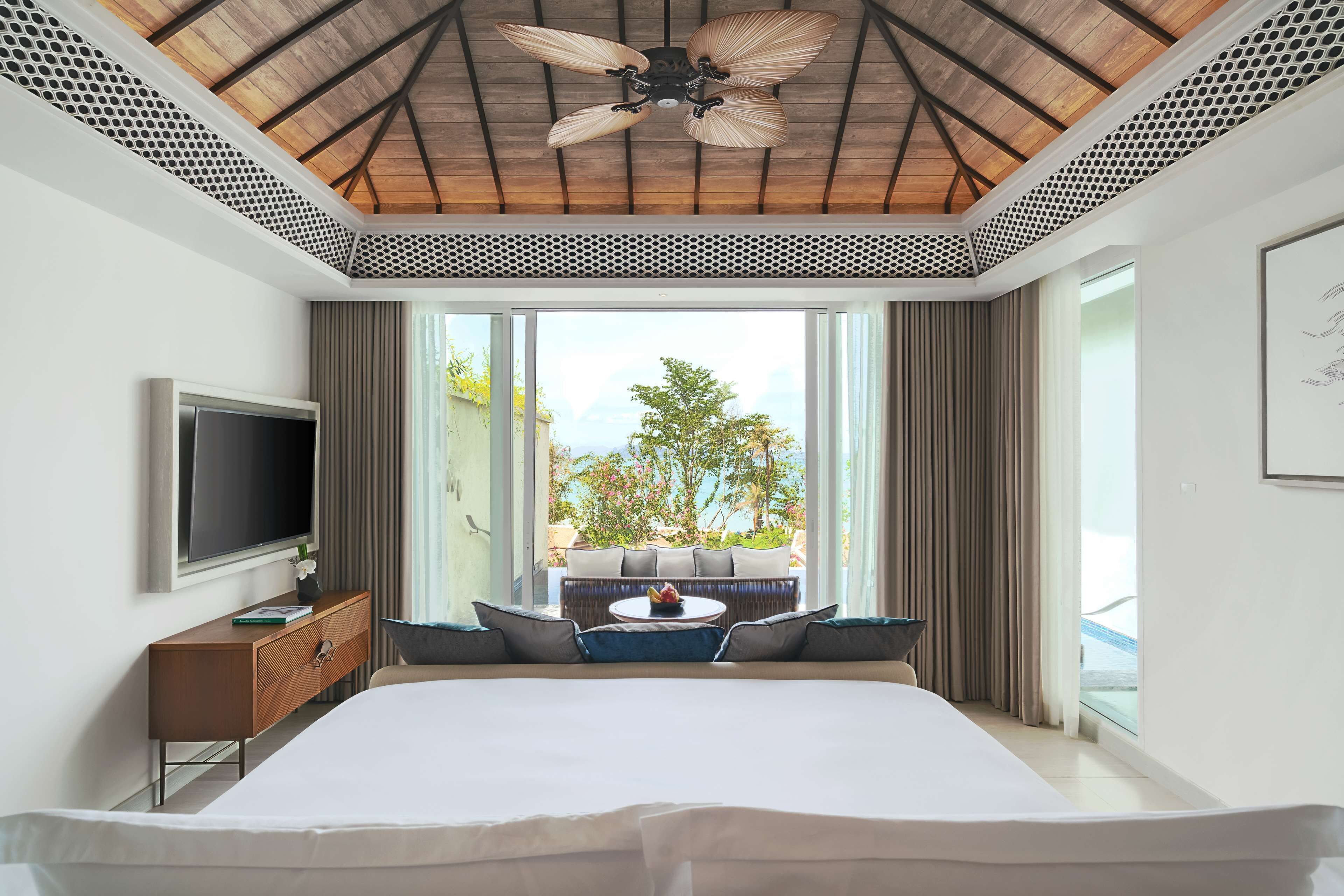 A modern hotel room with a large bed, a flat-screen TV, a wooden ceiling fan, and a balcony with a view of the outdoors.