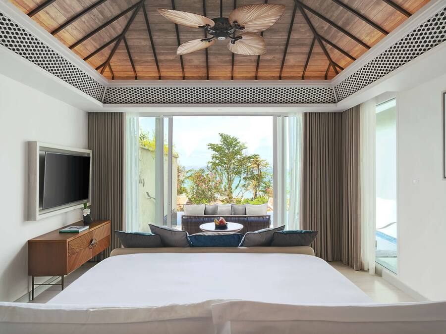 A modern hotel room with a large bed, a flat-screen TV, a wooden ceiling fan, and a balcony with a view of the outdoors.