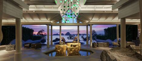 A modern interior with a central pool, seating areas, and a view of the sunset over water.