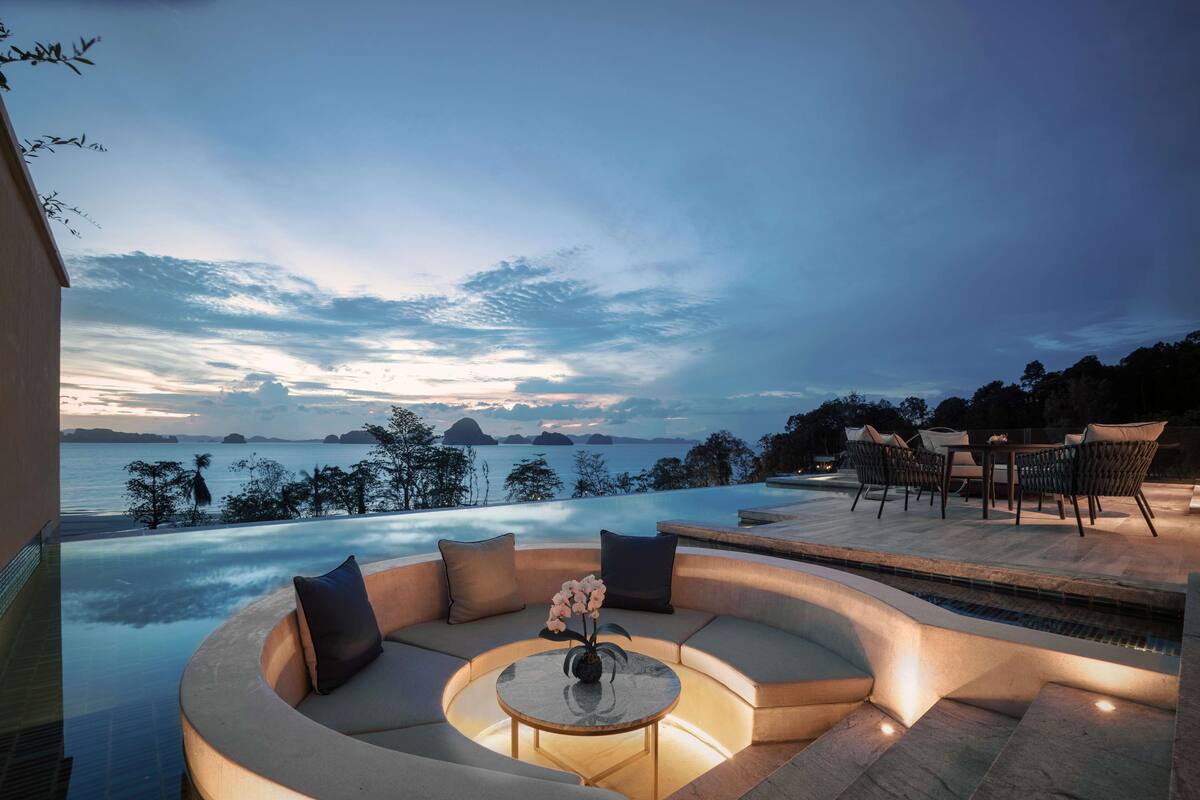 A luxurious outdoor seating area with a curved sofa and a round table, overlooking a large infinity pool and a scenic ocean view.