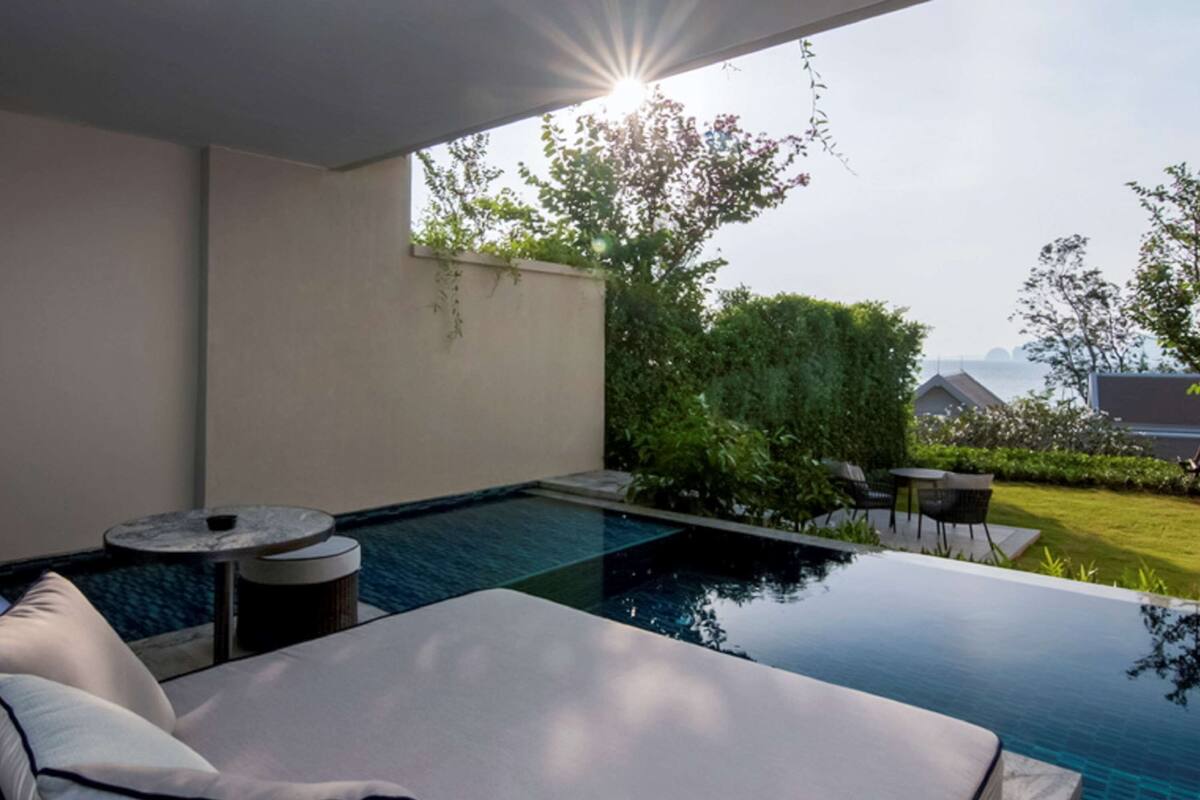 A modern outdoor pool area with a lounge chair and a small table, surrounded by greenery and a view of the landscape.