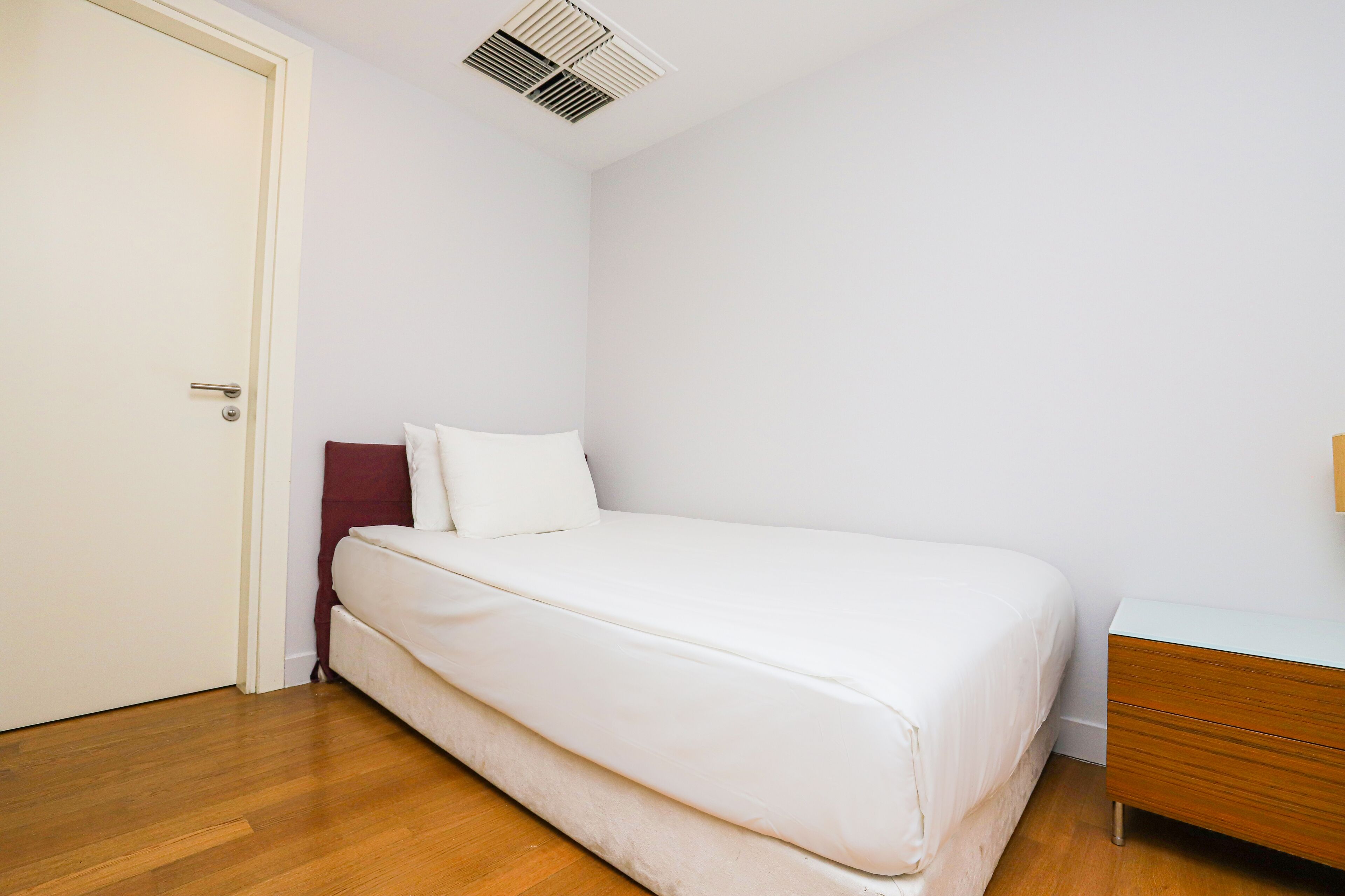 A single-bed room with a wooden floor, a white bed, a wooden nightstand, and a white door.