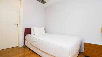 A single-bed room with a wooden floor, a white bed, a wooden nightstand, and a white door.
