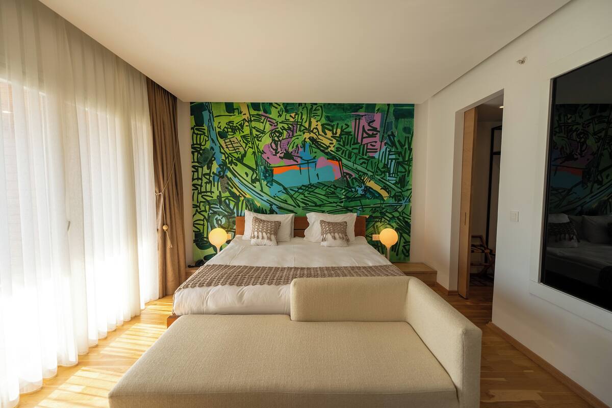Un dormitorio moderno con una cama grande, un sofá, un mural abstracto y colorido en la pared y un amplio ventanal con cortinas traslúcidas.