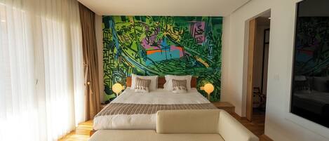 A modern bedroom with a large bed, a sofa, a colorful abstract mural on the wall, and a large window with sheer curtains.