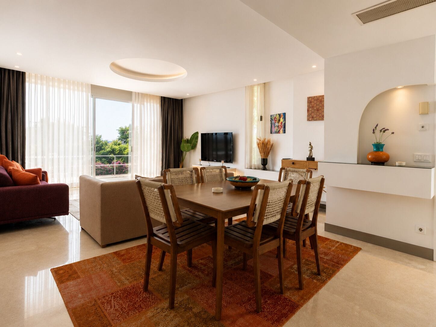 A modern living room with a sofa, a dining area with a table and chairs, a flat-screen TV, and a balcony with curtains.