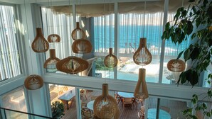 Amplio espacio interior con grandes ventanales con vista a una playa, que cuenta con varios rincones de descanso y lámparas colgantes de mimbre.