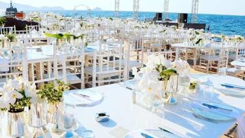 An outdoor wedding setup with white tables and chairs, arranged in rows, overlooking the sea.