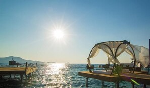 Un muelle iluminado por el sol con una marquesina y asientos, con vistas a aguas tranquilas y montañas lejanas.