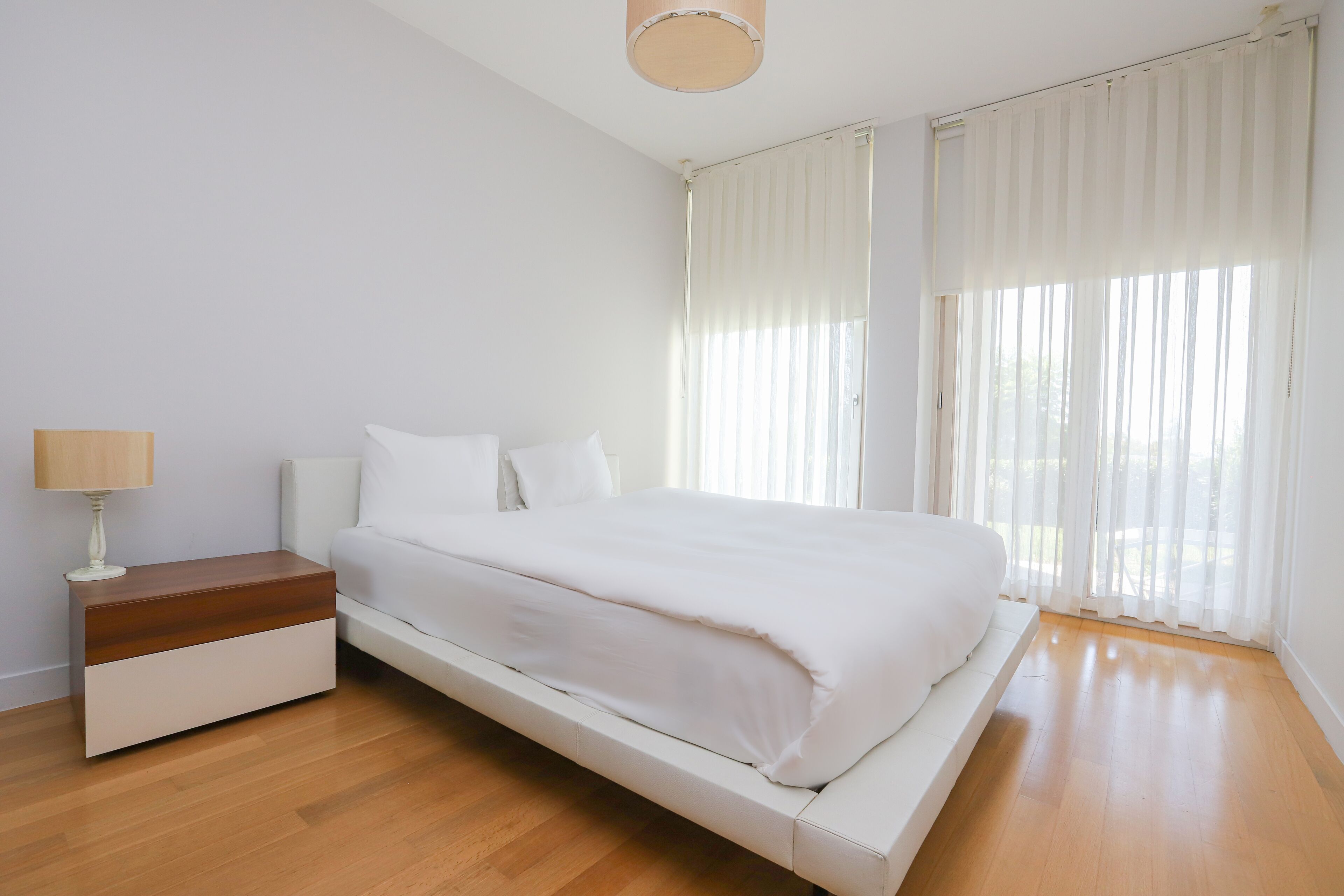 A modern bedroom with a bed, bedside table, and a window with sheer curtains.